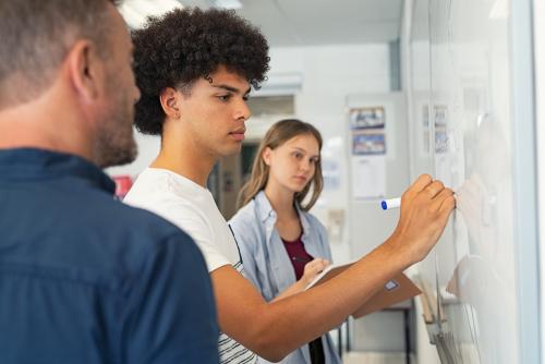 Two older students and their teacher standing at a whiteboard. One of the students is writing something.