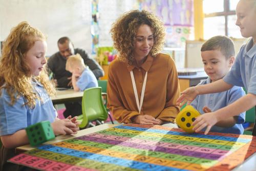 A teacher at a table with young children, looking at a colourful grid and dice.